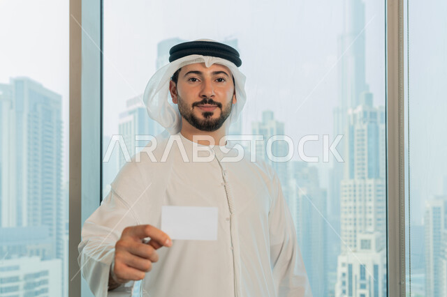 الوقوف باستقامة والنظر الى الكاميرا بايماءات السرور والسعادة، موك أب أبيض مستطيل فارغ، عرض بطاقة تعريفية، صورة مقربة لرجل عربي خليجي اماراتي يرتدي الكندورة والغترة البيضاء يحمل بيده هوية شخصية، بيئة العمل المريحة في شركات الامارات