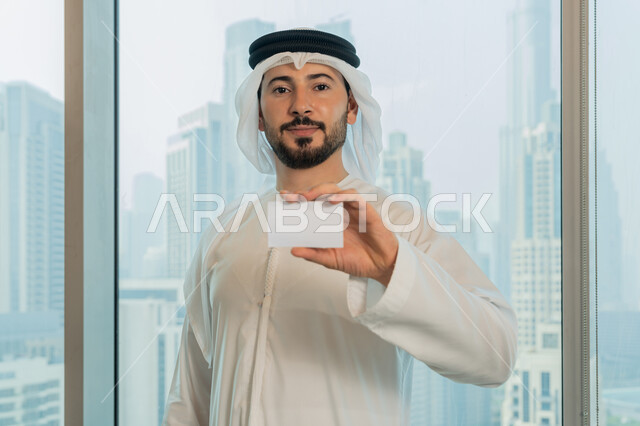 الوقوف باستقامة والنظر الى الكاميرا بايماءات السرور والسعادة، موك أب أبيض مستطيل فارغ، عرض بطاقة تعريفية، صورة مقربة لرجل عربي خليجي اماراتي يرتدي الكندورة والغترة البيضاء يحمل بيده هوية شخصية، بيئة العمل المريحة في شركات الامارات
