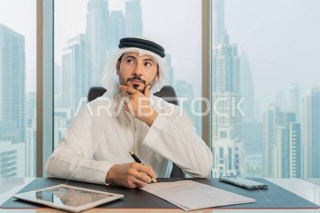تدوين وتسجيل الملاحظات والمعلومات على الورق, تنظيم وإدارة الأعمال، صورة مقربة لرجل عربي خليجي اماراتي يرتدي الكندورة والغترة البيضاء يقوم بكتابة المهام والأهداف اليومية بايماءات وجه ويدين تدل على التفكير والحيرة، التمعن والتركيز لايجاد حل المشكلة