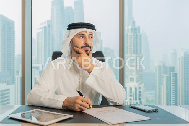 تدوين وتسجيل الملاحظات والمعلومات على الورق, تنظيم وإدارة الأعمال، صورة مقربة لرجل عربي خليجي اماراتي يرتدي الكندورة والغترة البيضاء يقوم بكتابة المهام والأهداف اليومية بايماءات وجه ويدين تدل على التفكير والحيرة، التمعن والتركيز لايجاد حل المشكلة