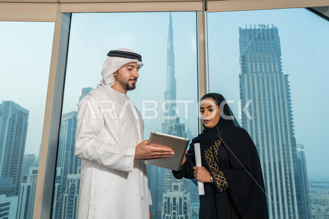 Using the tablet to explain the processes followed, understanding and cooperation between colleagues to achieve the company's goals, professionalism and dedication to achievement, an Arab Gulf Emirati man wearing a white kandura and ghutra holding a tablet in his hand explaining the work plan to his colleague, ensuring a clear and comprehensive understanding of the work