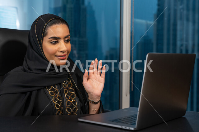 تصفح الانترنت وبرامج التواصل الاجتماعي, تطور التكنولوجيا وإستخدام الأجهزة التقنية الحديثة, صورة لامرأة عربية خليجية اماراتية محجبة ترتدي العباءة السوداء تجلس على الكرسي تقوم بالتواصل والقاء التحية مع العملاء, ادارة الأعمال من خلال جهاز الحاسوب المحمول