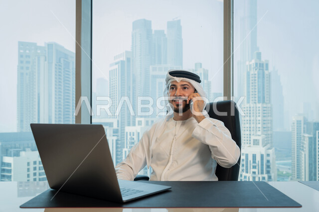 تصفح الانترنت وبرامج التواصل الاجتماعي, تطور التكنولوجيا وإستخدام الأجهزة التقنية الحديثة, صورة لرجل عربي خليجي اماراتي يرتدي الزي التقليدي ويقوم بإجراء مكالمة هاتفية عبر الجوال, ادارة الأعمال من خلال جهاز الحاسوب المحمول