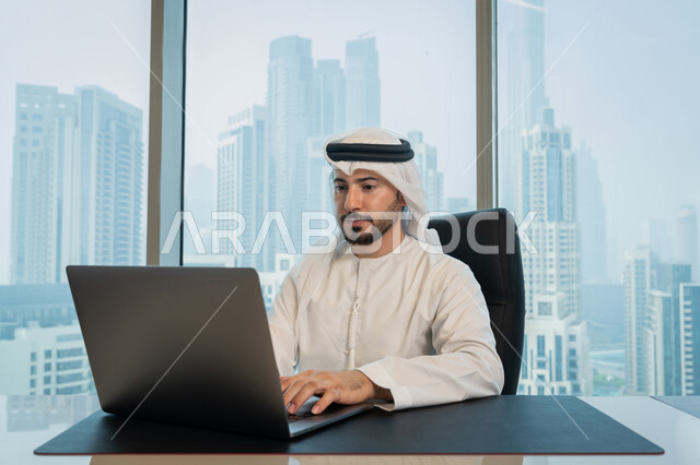 مهن ووظائف ادارية مكتبية, تصفح الانترنت وبرامج التواصل الاجتماعي, تطور التكنولوجيا وإستخدام الأجهزة التقنية الحديثة, صورة لرجل عربي خليجي اماراتي يرتدي الزي التقليدي ويقوم بادارة الأعمال من خلال جهاز الحاسوب المحمول