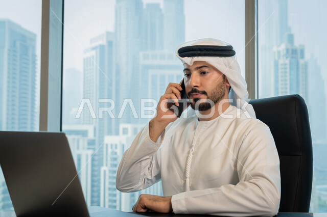 تصفح الانترنت وبرامج التواصل الاجتماعي, تطور التكنولوجيا وإستخدام الأجهزة التقنية الحديثة, صورة مقربة لرجل عربي خليجي اماراتي يرتدي الزي التقليدي ويقوم بإجراء مكالمة هاتفية عبر الجوال, ادارة الأعمال من خلال جهاز الحاسوب المحمول