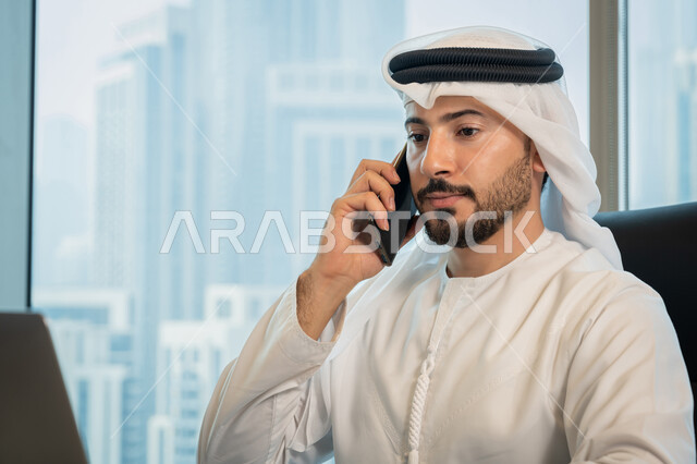 Browsing the internet and social networking programs, the development of technology and the use of modern technical devices, a close-up image of an Arab Gulf Emirati man wearing traditional clothing and making a phone call via mobile, business management through a laptop computer