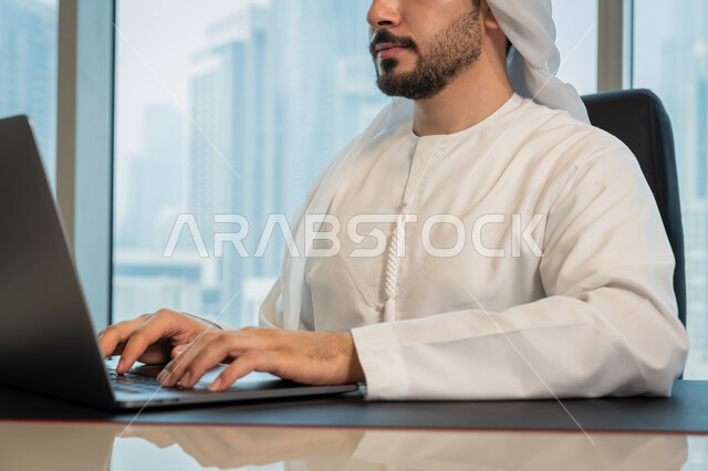 Data entry through the keyboard, the development of technology and the use of modern technical devices, a close-up image of an Arab Gulf Emirati man wearing traditional clothing and managing business through a laptop, browsing the Internet and social networking programs