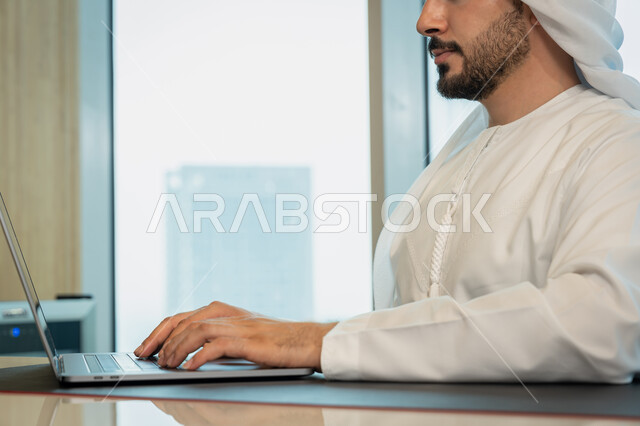 Data entry through the keyboard, the development of technology and the use of modern technical devices, a close-up image of an Arab Gulf Emirati man wearing traditional clothing and managing business through a laptop, browsing the Internet and social networking programs
