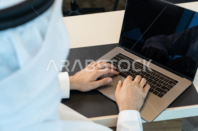 تطور التكنولوجيا وإستخدام الأجهزة التقنية الحديثة, صورة مقربة لرجل عربي خليجي اماراتي يرتدي الزي التقليدي ويقوم بادارة الأعمال من خلال الحاسوب المحمول، تصفح الانترنت وبرامج التواصل الاجتماعي، ادخال البيانات من خلال لوحة المفاتيح