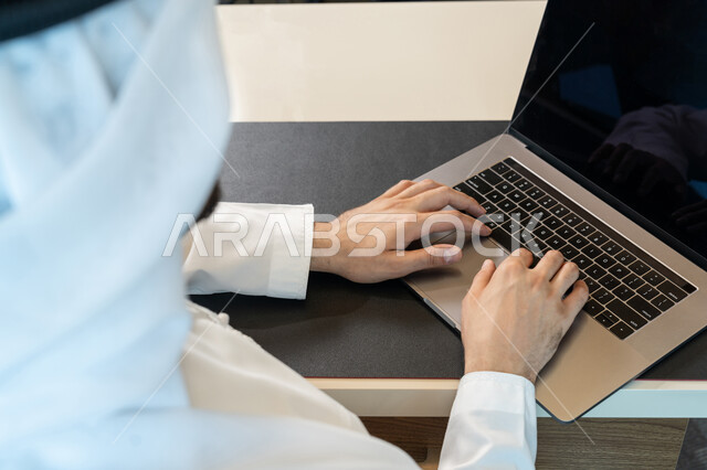 The development of technology and the use of modern technical devices, a close-up image of an Arab Gulf Emirati man wearing traditional clothing and managing business through a laptop, browsing the Internet and social networking programs, entering data through the keyboard
