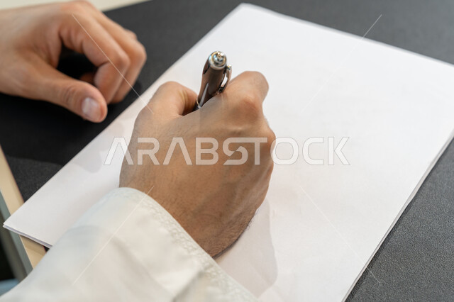 تدوين وتسجيل الملاحظات والمعلومات على الورق, صورة مقربة ليدين شاب عربي خليجي اماراتي يرتدي الثوب التقليدي يقوم بكتابة المهام والأهداف اليومية, تنظيم وإدارة الأعمال