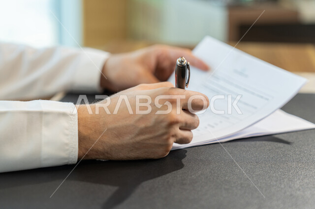 إدارة الأعمال وتنظيمها، تدوين وتسجيل الملاحظات والمعلومات على الورق, صورة مقربة ليدين شاب عربي خليجي اماراتي يرتدي الثوب التقليدي يقوم بكتابة المهام والأهداف اليومية