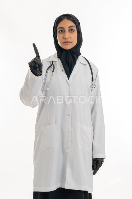 الأناقة والاهتمام بالمظهر الخارجي،  مفهوم الطب والرعاية الصحية, العمل في المراكز الصحية والمستشفيات، تقديم المساعده الطبية للمرضى, بورتريه لطبيبة عربية خليجية اماراتية محجبة ترتدي المعطف الطبي الأبيض وسماعة الفحص وترفع اصبع السبابة للاعلى, خلفية بيضاء
