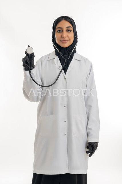الطب والرعاية الصحية، مفهوم الأنوثة والنعومة, العمل في المراكز الصحية والمستشفيات، تقديم المساعده الطبية للمرضى, بورتريه لطبيبة عربية خليجية اماراتية محجبة ترتدي المعطف الطبي الأبيض وترفع بيدها سماعة الفحص الطبية , خلفية بيضاء