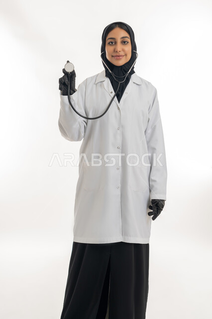 Medicine and health care, the concept of femininity and softness, working in health centers and hospitals, providing medical assistance to patients, portrait of a veiled Gulf Arab Emirati female doctor wearing a white medical coat and raising a stethoscope in her hand, white background