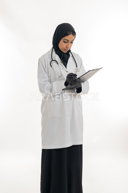 العمل في القطاع الصحي, مفهوم الأنوثة والنعومة، تقديم المساعده الطبية للمرضى, بورتريه لطبيبة عربية خليجية اماراتية محجبة ترتدي المعطف الطبي الأبيض تقوم بإعداد التقارير الطبية, تشخيص الحالات المرضية, خلفية بيضاء