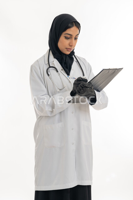 Elegance and interest in appearance, the concept of femininity and softness, providing assistance to patients, portrait of a veiled female Gulf Arab Emirati doctor wearing a white medical coat preparing medical reports, diagnosing medical cases, white background