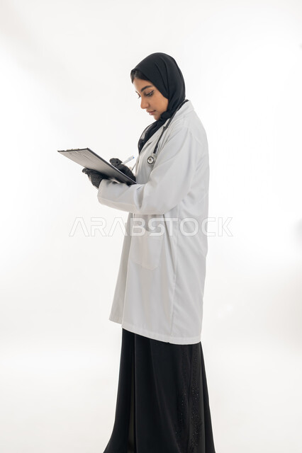 الأناقة والاهتمام بالمظهر الخارجي، مفهوم الأنوثة والنعومة، تقديم المساعده للمرضى, بورتريه لطبيبة عربية خليجية اماراتية محجبة ترتدي المعطف الطبي الأبيض تقوم بإعداد التقارير الطبية, تشخيص الحالات المرضية, خلفية بيضاء