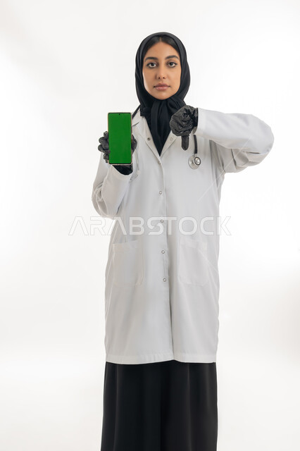 الطب والرعاية الصحية، مفهوم الأنوثة والنعومة، تقديم المساعده للمرضى, بورتريه لطبيبة عربية خليجية اماراتية محجبة ترتدي المعطف الطبي الأبيض تشير الى هاتف محمول بشاشة كروما خضراء بإيماءات يدين تدل على عدم الاعجاب، خلفية بيضاء