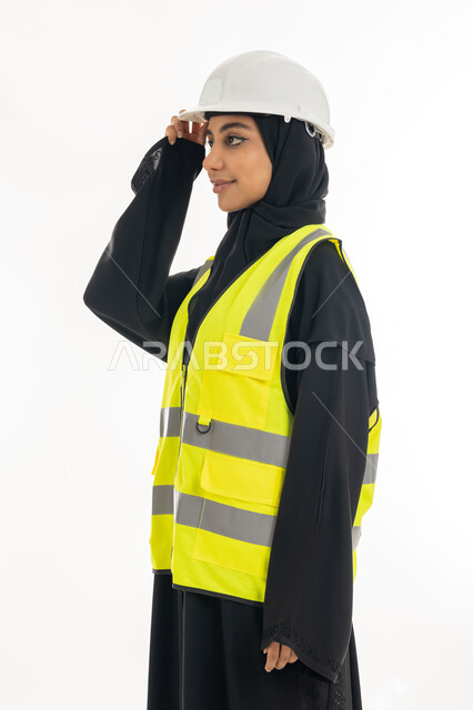 Women's jobs and professions, standing with confident gestures, the concept of engineering project management, working in the engineering sector, a portrait from the side of an Arab Gulf Emirati female architect wearing a protective helmet and jacket looking somewhere, white background