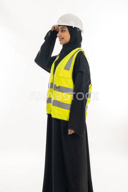 Standing with confident gestures, the concept of engineering project management, women’s jobs and professions, working in the engineering sector, a portrait from the side of an Arab Gulf Emirati female architect wearing a protection helmet and jacket looking somewhere, white background