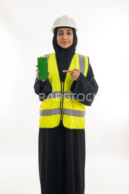 مفهوم ادارة المشاريع الهندسية, وظائف ومهن نسائية, دمج العمل مع التكنولوجيا، العمل في القطاع الهندسي, بورتريه لمهندسة معمارية عربية خليجية امارتية ترتدي خوذة الحماية والسترة تقوم بعرض هاتف محمول بشاشة خضراء، خلفية بيضاء