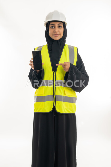 وظائف ومهن نسائية, دمج العمل مع التكنولوجيا، مفهوم ادارة المشاريع الهندسية,  العمل في القطاع الهندسي, بورتريه لمهندسة معمارية عربية خليجية امارتية ترتدي خوذة الحماية وتقوم بعرض هاتف محمول بشاشة سوداء، خلفية بيضاء