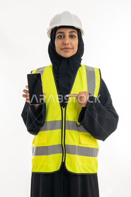 Integrating work with technology, the concept of engineering project management, women’s jobs and professions, working in the engineering sector, portrait of an Arab Gulf Emirati female architect wearing a protection helmet and displaying a mobile phone with a black screen, white background