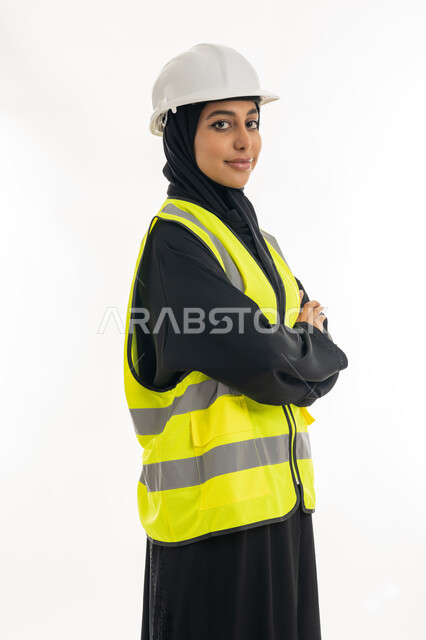 Standing with gestures of confidence, the concept of engineering project management, women’s jobs and professions, working in the engineering sector, portrait of an Arab Gulf Emirati female architect wearing a protective helmet and jacket and standing with crossed hands, white background