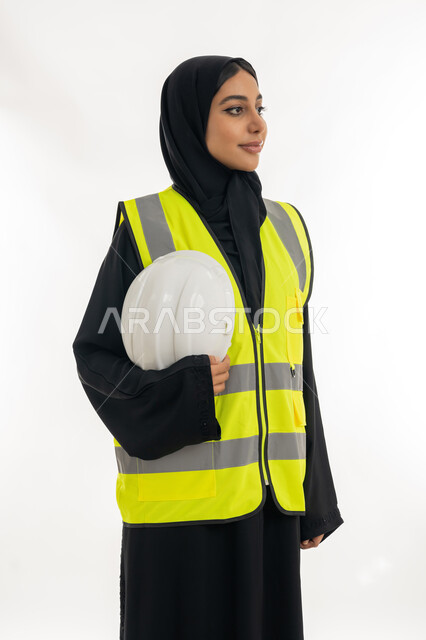 مفهوم الهندسة وأعمال البناء, مهن ووظائف نسائية، تقدم وتطور الامارات على يد أبناء الوطن، شابة سعودية تمسك بيدها خوذة الحماية، بورتريه لمهندسة عربية خليجية اماراتية محجبة ترتدي عباءة سوداء مع السترة تعمل في القطاع الهندسي الاماراتي, خلفية بيضاء