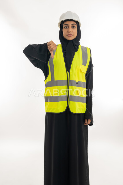 Feeling of rejection and abstention, progress and development of the Emirates by the sons of the homeland, the concept of engineering and construction work, a young Saudi woman raising her thumb up with a gesture of annoyance, a portrait of a veiled Arab Gulf Emirati engineer wearing a jacket and a protective helmet working in the engineering sector, white background
