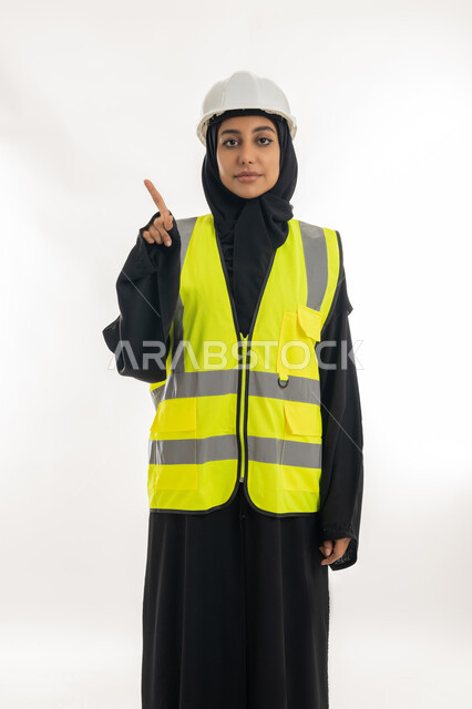 تقدم وتطور الامارات على يد أبناء الوطن، مهن ووظائف نسائية، مفهوم الهندسة وأعمال البناء, شابة سعودية تشير باصبع السبابة الى مكان ما, بورتريه لمهندسة عربية خليجية اماراتية محجبة ترتدي عباءة سوداء مع السترة وخوذة الحماية تعمل في القطاع الهندسي, خلفية بيضاء