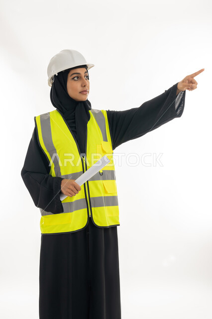 مهن ووظائف نسائية، مفهوم الهندسة وأعمال البناء, شابة سعودية تشير باصبع السبابة الى مكان ما, بورتريه لمهندسة عربية خليجية اماراتية محجبة ترتدي عباءة سوداء مع السترة وخوذة الحماية تمسك بيدها المخططات الهندسيه، العمل في القطاع الهندسي في الامارات, خلفية بيضاء
