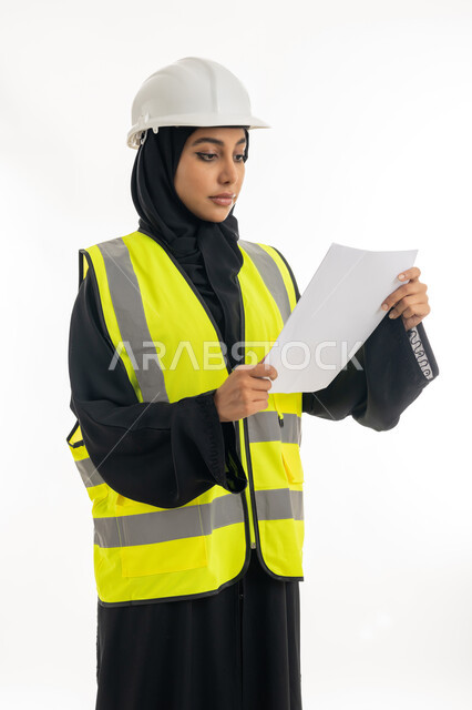 مفهوم الهندسة وأعمال البناء, شابة سعودية تمسك بيدها أوراق الملاحظات, بورتريه لمهندسة عربية خليجية اماراتية محجبة ترتدي عباءة سوداء مع السترة وخوذة الحماية تقوم بدراسه وتصفح المخططات الهندسيه، مهن ووظائف نسائية، العمل في القطاع الهندسي في الامارات, خلفية بيضاء