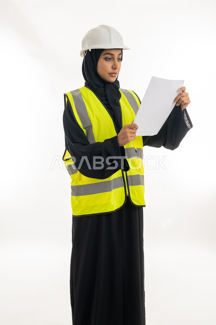 مفهوم الهندسة وأعمال البناء, شابة سعودية تمسك بيدها أوراق الملاحظات, بورتريه لمهندسة عربية خليجية اماراتية محجبة ترتدي عباءة سوداء مع السترة وخوذة الحماية تقوم بدراسه وتصفح المخططات الهندسيه، مهن ووظائف نسائية، العمل في القطاع الهندسي في الامارات, خلفية بيضاء