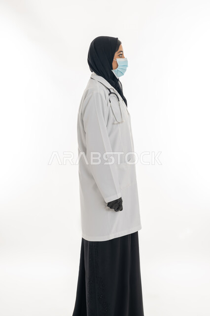 الالتزام بالاحترازات الوقائيه ضد كوفيد 19، العمل في القطاع الصحي الاماراتي, مهن ووظائف نسائية، شابة سعودية تقدم الرعاية الطبية للمرضى، بورتريه لطبيبة عربية خليجية اماراتية محجبة ترتدي العباءة السوداء مع المعطف الطبي الأبيض وكمامة الوقاية تقف باستقامة، خلفية بيضاء