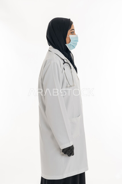 Adherence to preventive precautions against Covid-19, working in the UAE health sector, women's professions and jobs, a young Saudi woman providing medical care to patients, portrait of a veiled Emirati Gulf Arab female doctor wearing a black abaya with a white medical coat and a protective mask standing straight, white background