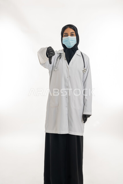 Feeling of rejection and abstention, a young Saudi woman providing medical care to patients, portrait of an Arab Gulf Emirati female doctor wearing a white medical coat and a protective mask pointing her thumb down with gestures of annoyance and negative evaluation, commitment to preventive precautions against Covid-19, white background