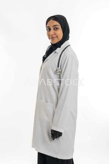تقديم المساعده الطبية للمرضى, مهن ووظائف نسائية,  شابة سعودية تقوم بالعمل في المراكز والمستشفيات, بورتريه من الجانب لطبيبة عربية خليجية اماراتية ترتدي المعطف الطبي الأبيض وسماعة الفحص تنظر إلى الكاميرا بايماءات الثقة بالنفس, مفهوم الطب والرعاية الصحية, خلفية بيضاء