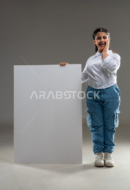 استخدام اللوحات التوضيحيه للعرض والتقديم, مفهوم الاعلان والتسويق، بورتريه لطفلة عربية خليجية عمانية ترتدي الزي الكاجوال تقف بجانب لوحة بيضاء كبيرة،  لوحة فنية فارغة، موك اب أبيض، صورة الجسم بالطول الكامل، خلفية رمادية