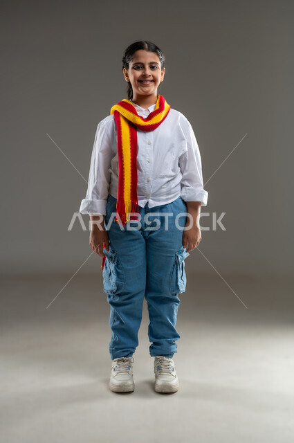 ارتداء اوشحة تشجيعية للفريق المفضل، الشغف في متابعة الأندية الرياضية منذ الصغر،  تشجيع الفريق العماني، بورتريه لفتاة عربية خليجية سعودية مبتسمة ترتدي الزي الكاجوال تنظر الى الكاميرا بسعادة، الحماس لتحقيق الفوز و الانتصار، صورة الجسم بالطول الكامل, خلفية رمادية