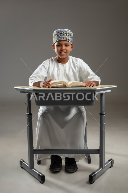 المناهج العلمية التعليمية في سلطنة عمان، ايماءات الاندماج والتركيز, مفهوم حب القراءة والمطالعة, الجد والاجتهاد للوصول إلى النجاح، العودة الى المدارس، بورتريه  لطالب عربي عماني  يرتدي الدشداشة والكمة يجلس على مقعد ينظر الى الكاميرا بابتسامة، خلفية رمادية