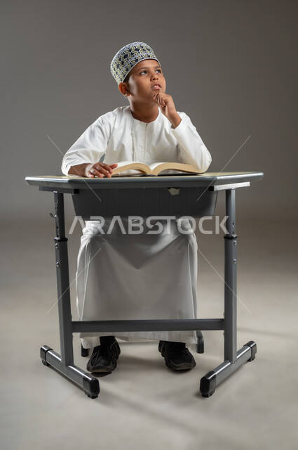 المناهج العلمية التعليمية في سلطنة عمان، ايماءات الاندماج والتركيز, مفهوم حب القراءة والمطالعة, الجد والاجتهاد للوصول إلى النجاح، العودة الى المدارس، بورتريه  لطالب عربي عماني  يرتدي الدشداشة والكمة يجلس على مقعد ينظر الى مكان ما بايماءات التفكير، خلفية رمادية