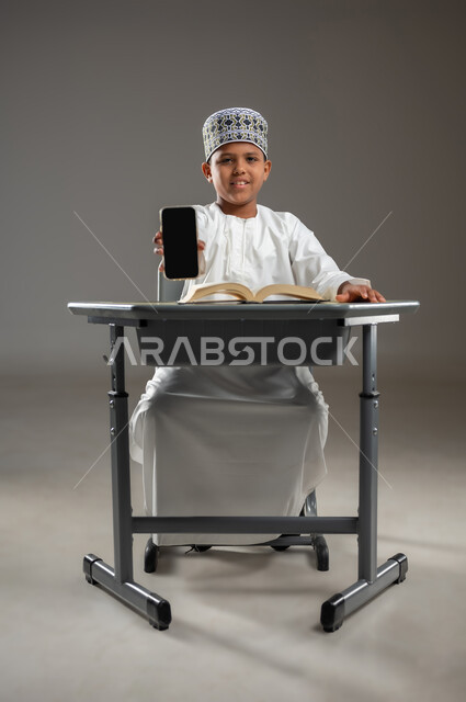 عرض خدمات وتطبيقات الهاتف المحمول, الجد والاجتهاد للوصول إلى النجاح، مفهوم حب القراءة والمطالعة, العودة الى المدارس، بورتريه جانبي لطالب عربي عماني  يرتدي الدشداشة والكمة يقوم بعرض شاشة سوداء فارغة، المناهج العلمية التعليمية في سلطنة عمان، خلفية رمادية