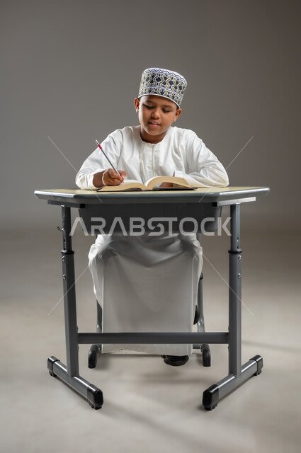 Back to school, gestures of integration and focus, concept of love of reading and studying, seriousness and diligence to achieve success, portrait of an Arab Omani student wearing a dishdasha and a cloak sitting on a chair holding a book and a pencil, educational scientific curricula in the Sultanate of Oman, gray background