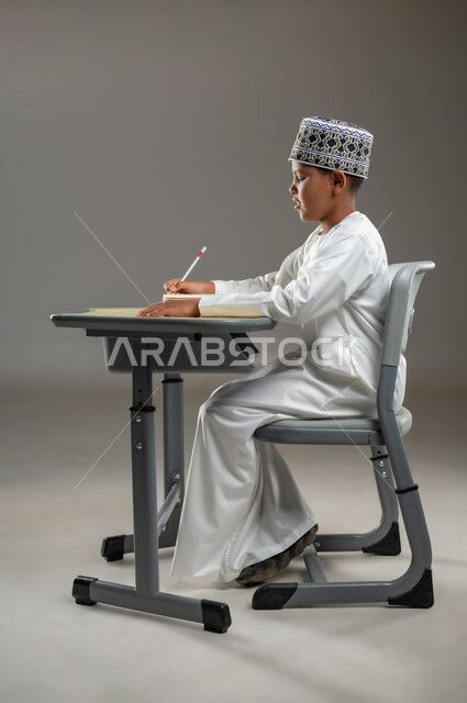 الجد والاجتهاد للوصول إلى النجاح، ايماءات الاندماج والتركيز, مفهوم حب القراءة والمطالعة, العودة الى المدارس، بورتريه جانبي لطالب عربي عماني  يرتدي الدشداشة والكمة يجلس على مقعد يمسك بيده كتاب وقلم رصاص، المناهج العلمية التعليمية في سلطنة عمان، خلفية رمادية