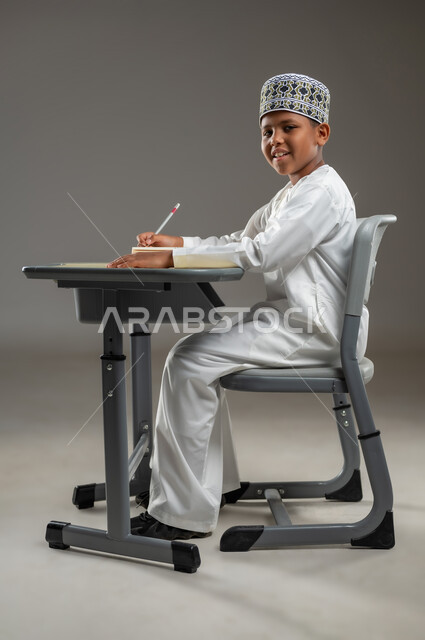 ايماءات الاندماج والتركيز, مفهوم حب القراءة والمطالعة, الجد والاجتهاد للوصول إلى النجاح، العودة الى المدارس، بورتريه جانبي لطالب عربي عماني  يرتدي الدشداشة والكمة يجلس على مقعد يمسك بيده كتاب وقلم رصاص، المناهج العلمية التعليمية في سلطنة عمان، خلفية بيضاء