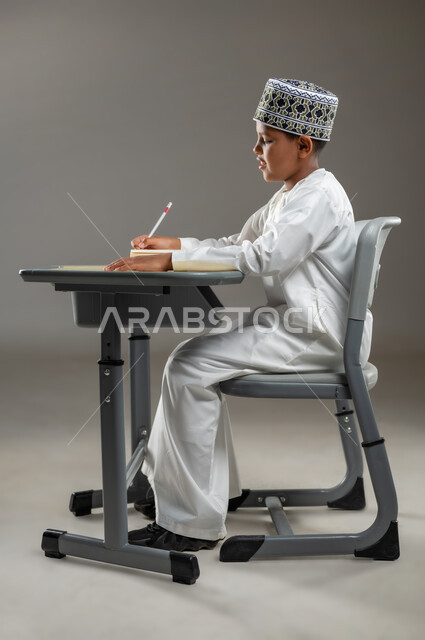 ايماءات الاندماج والتركيز, مفهوم حب القراءة والمطالعة, الجد والاجتهاد للوصول إلى النجاح، العودة الى المدارس، بورتريه جانبي لطالب عربي عماني  يرتدي الدشداشة والكمة يجلس على مقعد يمسك بيده كتاب وقلم رصاص، المناهج العلمية التعليمية في سلطنة عمان، خلفية بيضاء