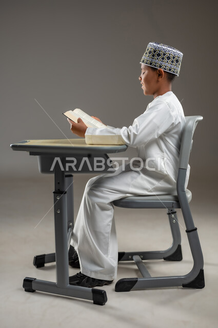 مفهوم حب القراءة والمطالعة, الجد والاجتهاد للوصول إلى النجاح، العودة الى المدارس، بورتريه لطالب عربي خليجي عماني  يرتدي الدشداشة والكمة يجلس على مقعد يمسك بيده الكتاب، المناهج العلمية التعليمية في سلطنة عمان، خلفية بيضاء