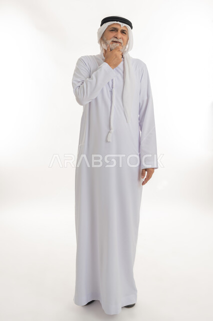 إيماءات التفكير العميق والحيرة، وضع اليد تحت الذقن بتعابير القلق وعدم اتخاذ القرار، بورتريه لرجل عربي خليجي إماراتي كبير بالسن يرتدي الكندورة الإماراتية التقليدية والغترة يقف وينظر إلى مكان بعيد ويخطط لشيء ما، الجسم بالطول الكامل، خلفية بيضاء